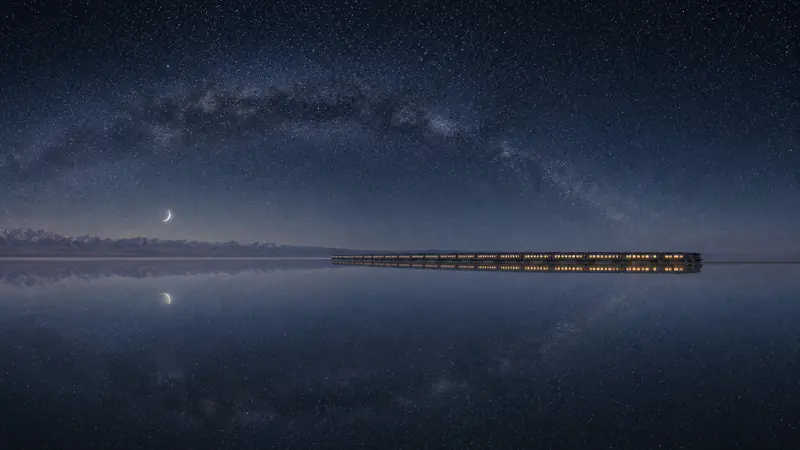 From far away, the tiny lit train crosses the vast salt flat under the arc of the Milky Way, the whole sky reflected below it.